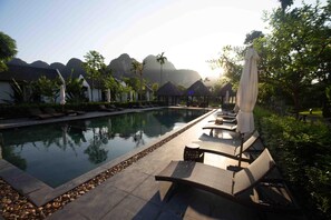 A pool area with lounge chairs and umbrellas, surrounded by trees and buildings with thatched roofs. - Aravinda Resort (Nam Hoa Lu)