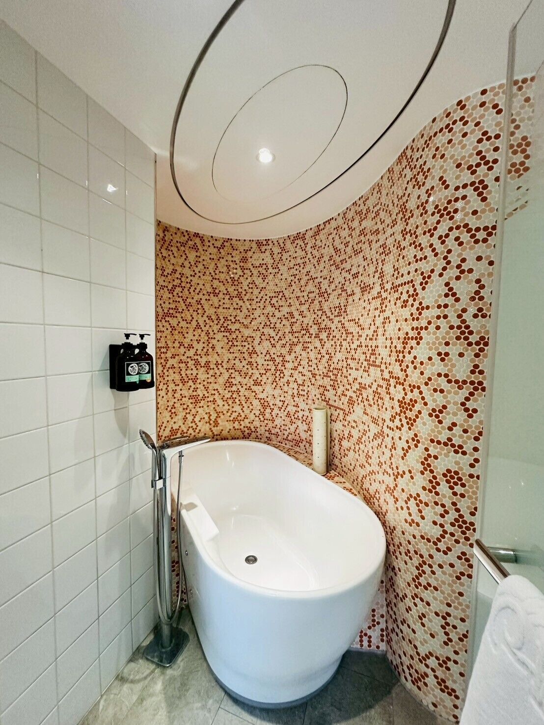 A modern bathroom with a freestanding bathtub, white tiled walls, and a patterned mosaic wall.