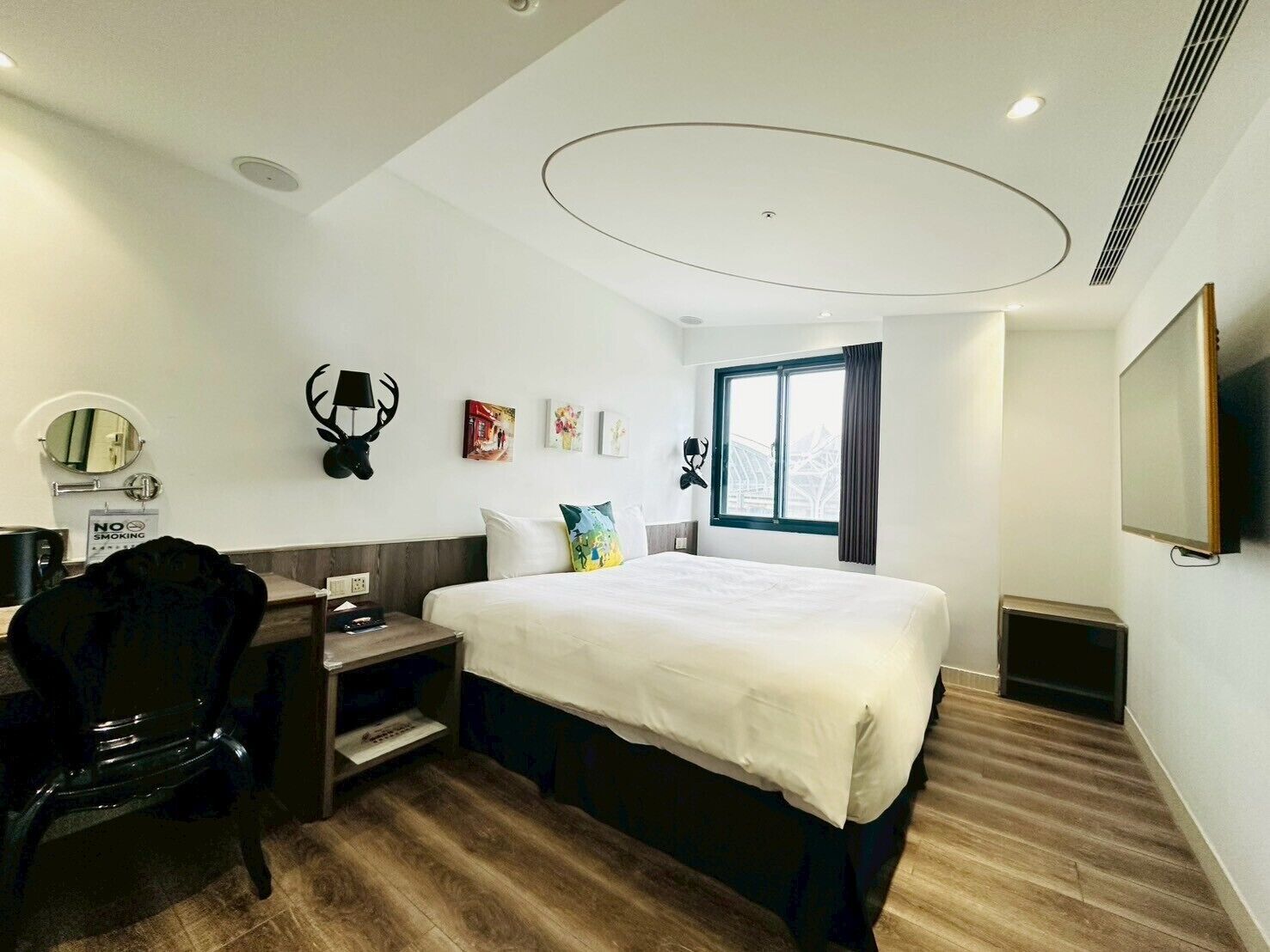 A hotel room with a large bed, a desk with a chair, a mounted deer head, and a window with curtains.