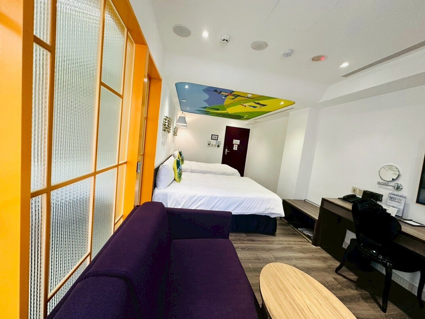 A hotel room with two beds, a desk, a chair, a sofa, and a large window with blinds.