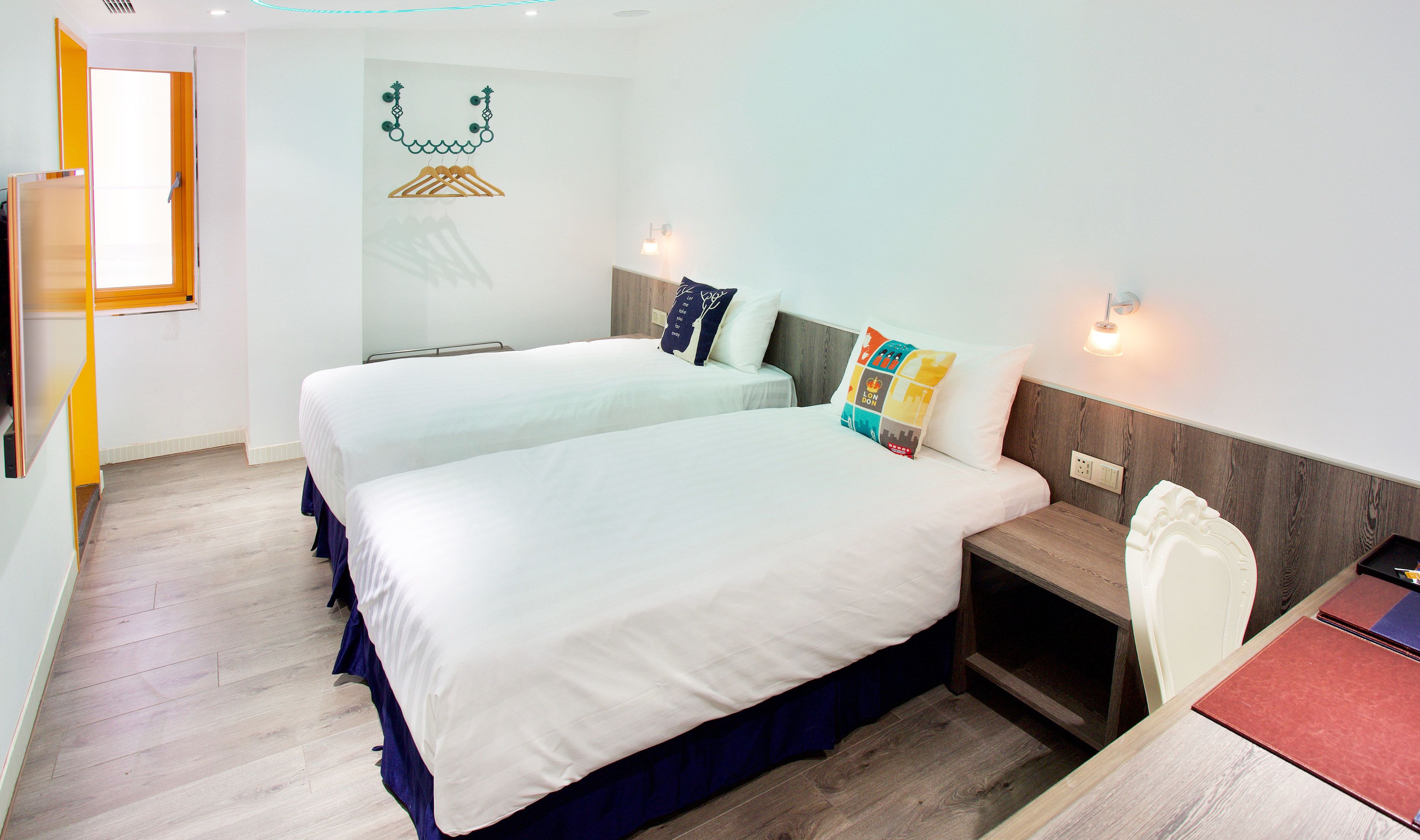 Una habitación de hotel con dos camas, suelo de madera, una mesita de noche y una lámpara fijada a la pared.