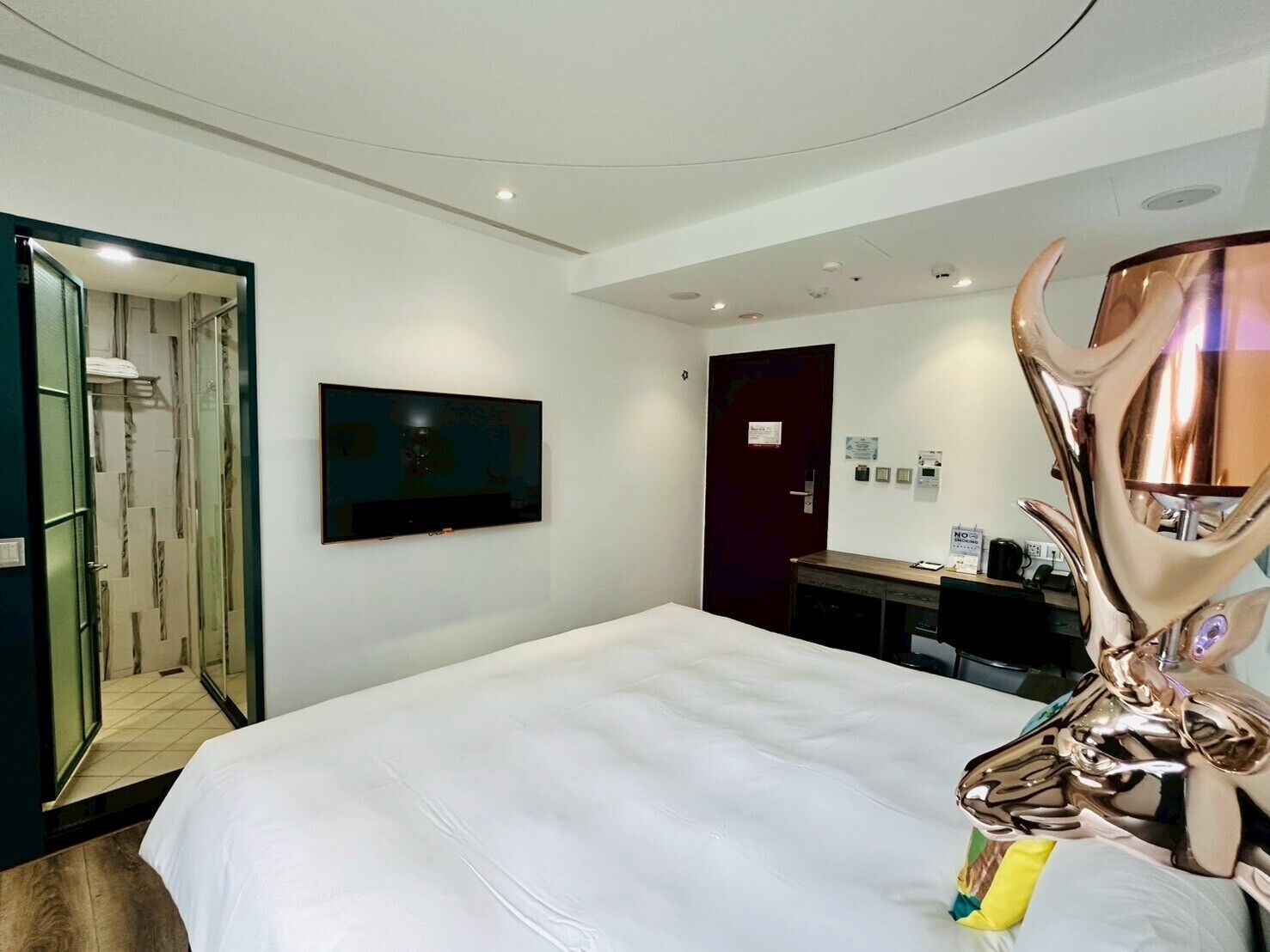 Una habitación de hotel con una cama, televisión montada en la pared, una puerta y una escultura decorativa.