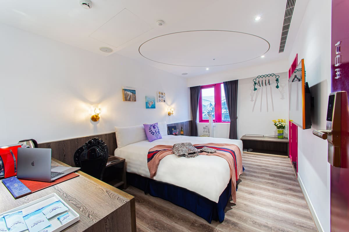 A modern hotel room with a large bed, a desk with a laptop, and a view of the city through the window.