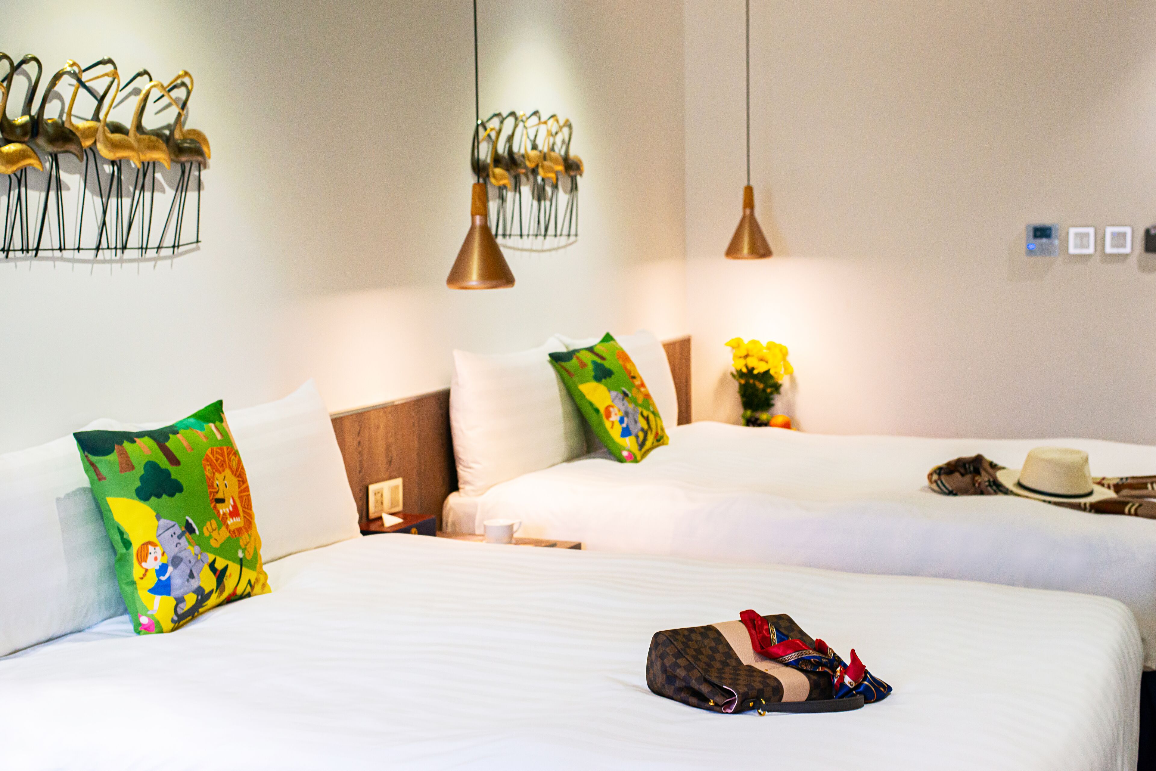 A hotel room with two beds, white bedding, colorful pillows, a small table with a cup, and a hat on the bed.