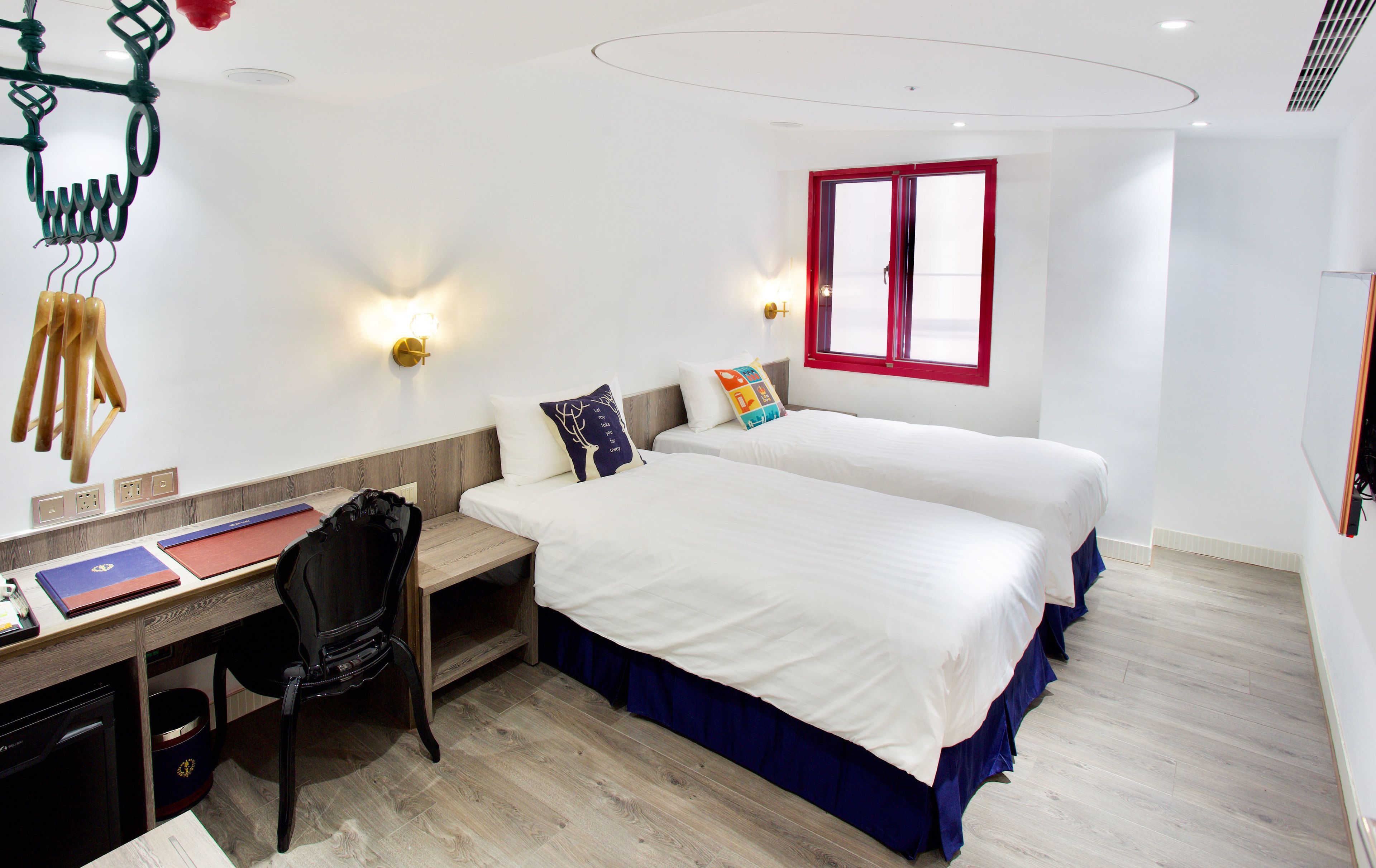 Quarto de hotel com duas camas, uma escrivaninha com cadeira, uma mesinha e uma janela com moldura vermelha.
