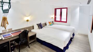 Quarto de hotel com duas camas, uma escrivaninha com cadeira, uma mesinha e uma janela com moldura vermelha.