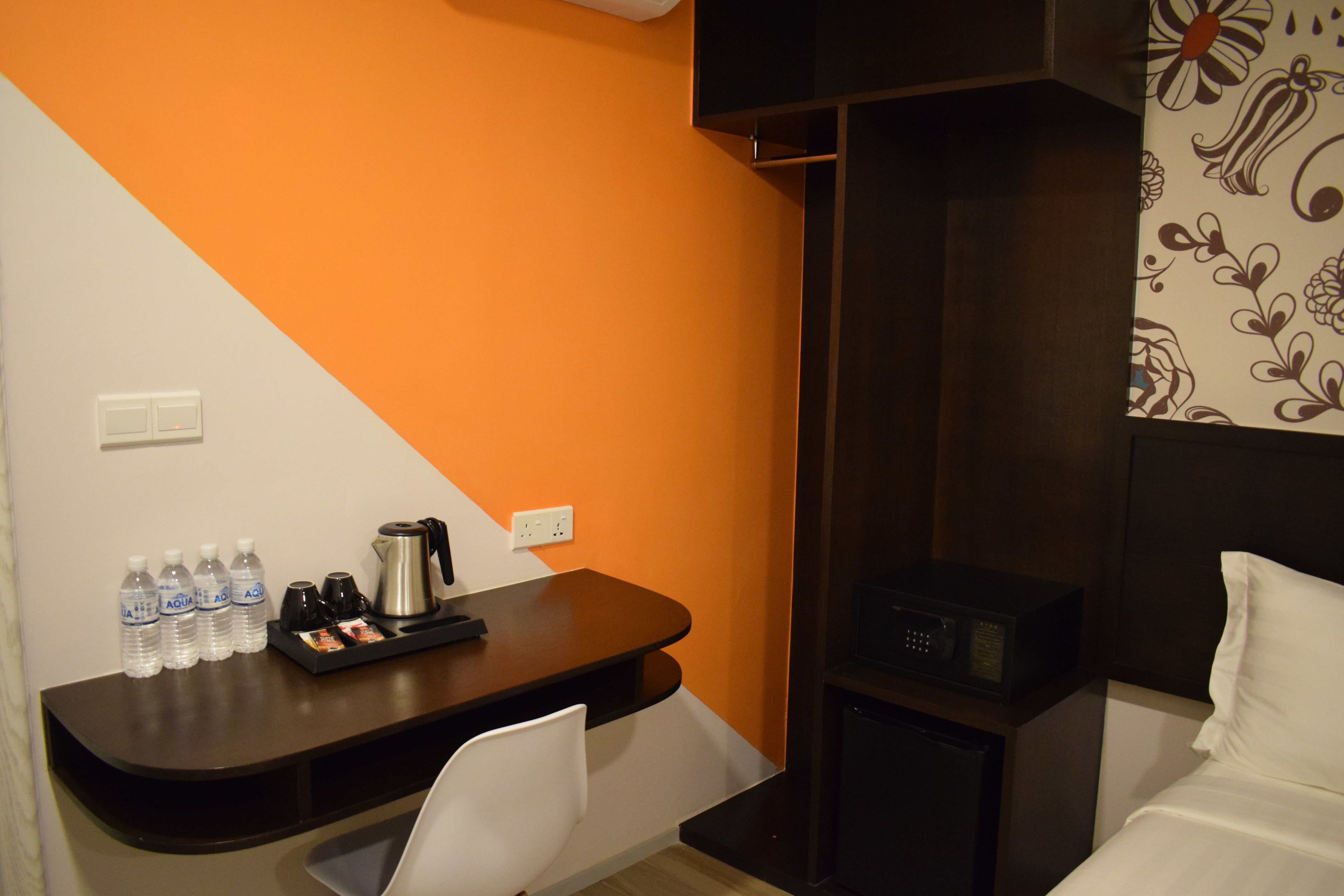 Habitación de hotel con un escritorio marrón, una silla blanca y una cama con ropa blanca. Hay botellas de agua, una tetera y un microondas sobre el escritorio.