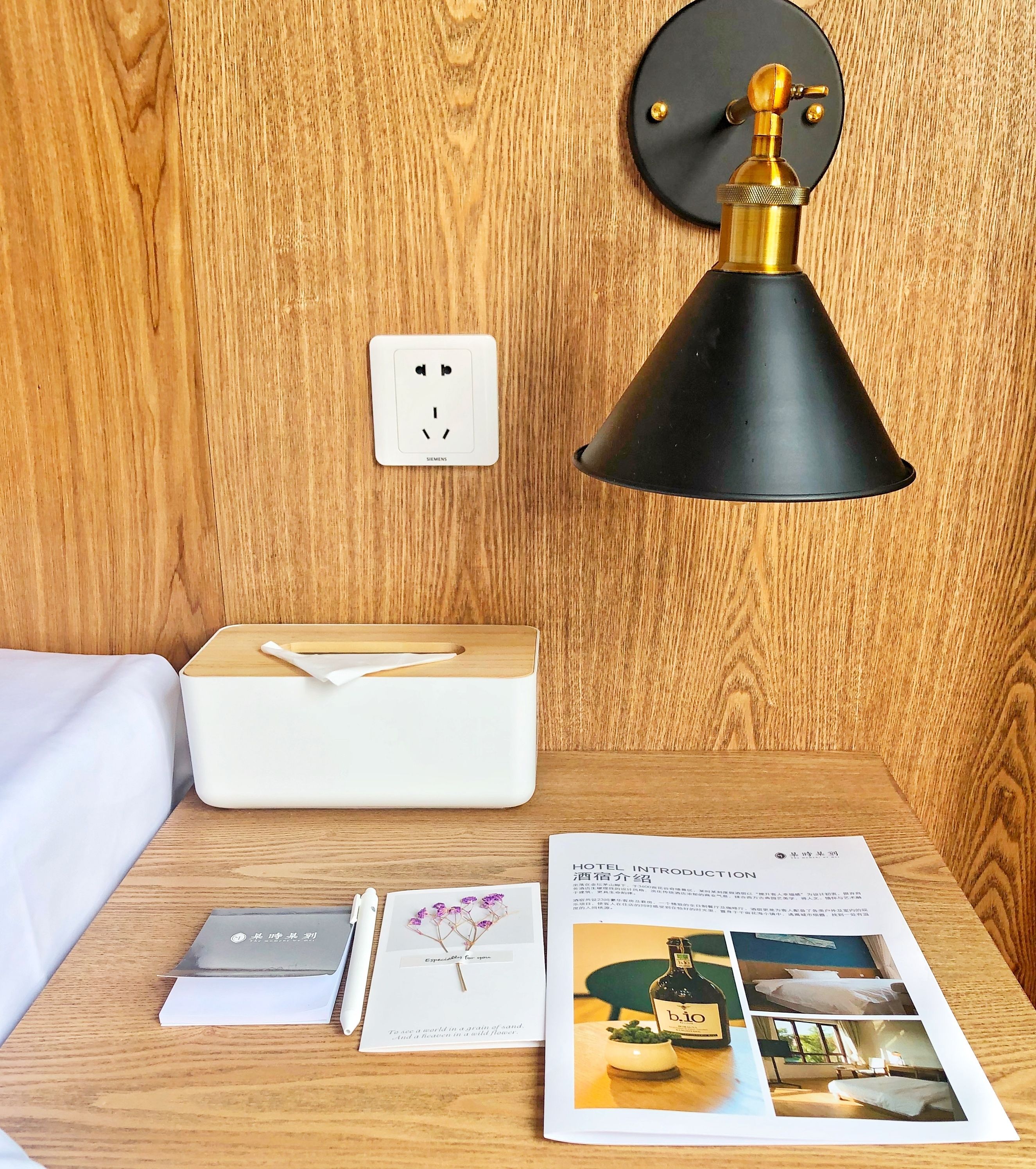 A neatly made bed with a white pillow and a wooden bedside table. On the table, there is a tissue box, a pen, a booklet, and a brochure.