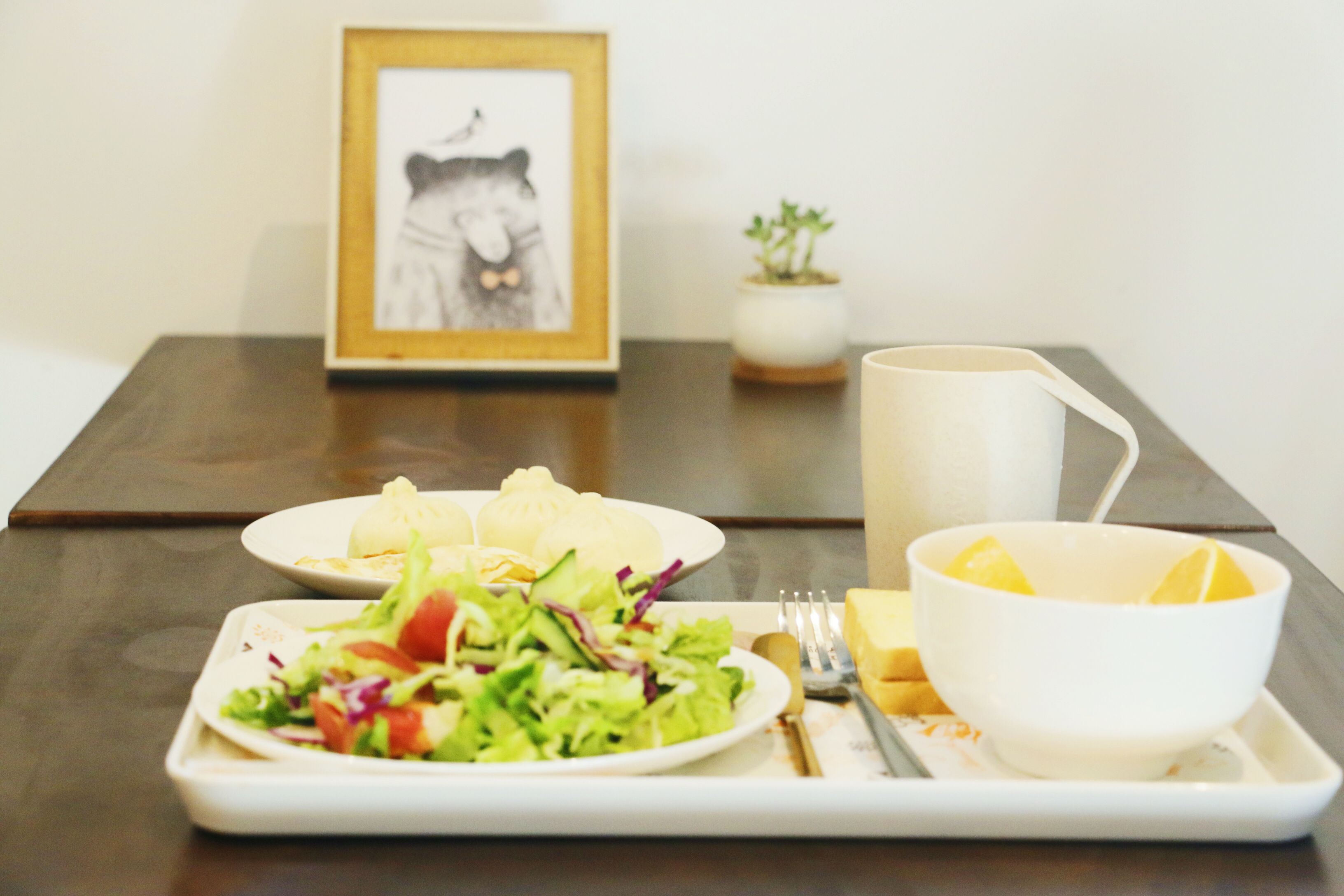Una mesa de comedor con un plato de comida, un bol de fruta, un cuadro enmarcado y una planta pequeña.