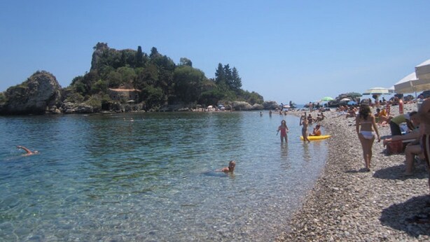 Ein Kiesstrand mit klarem Wasser, Menschen beim Schwimmen und einer felsigen Insel im Hintergrund.