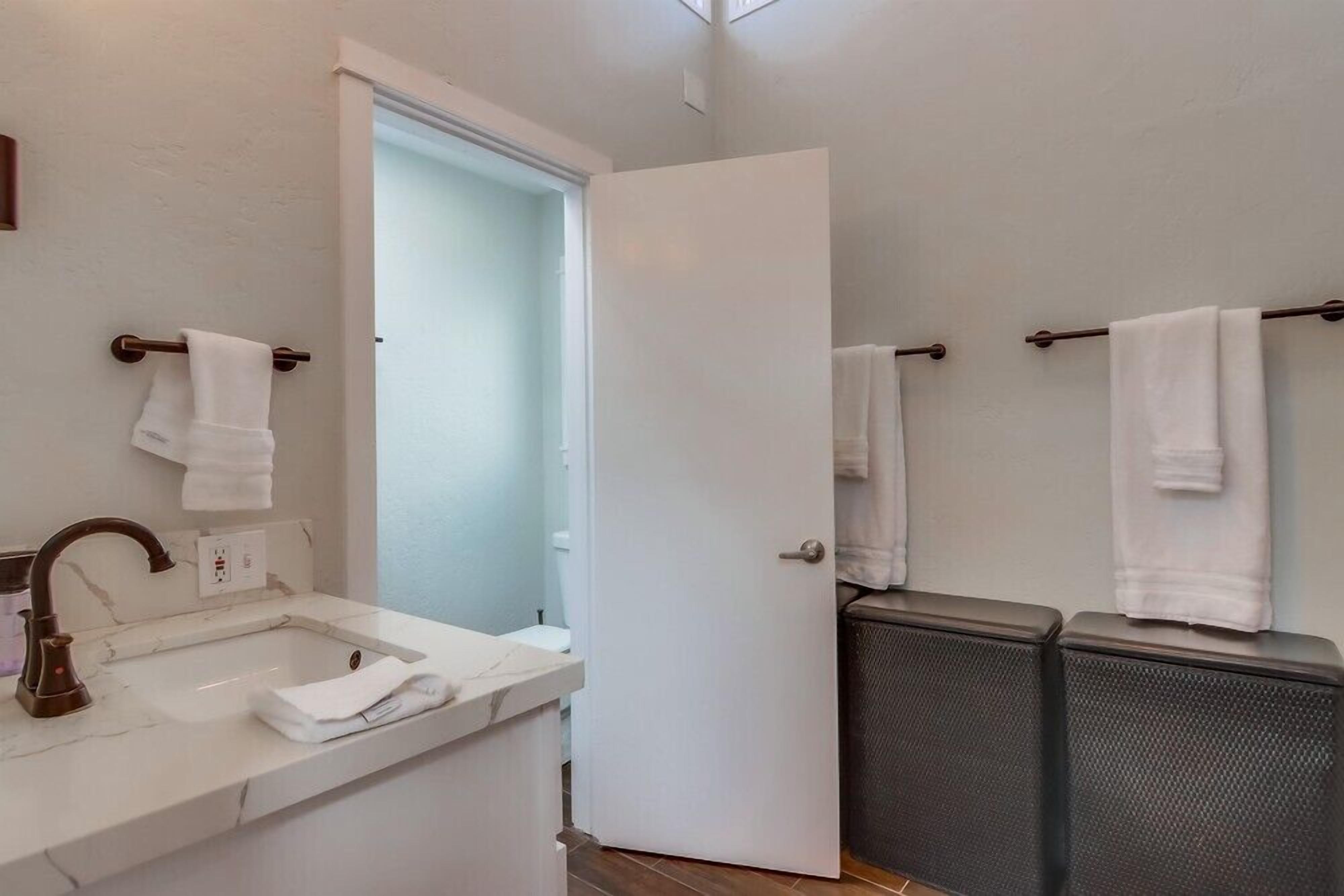 Un baño con lavamanos, espejo, toallero y una puerta que da a otra habitación.
