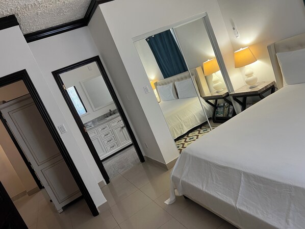 A hotel room with a bed, a desk, a chair, a mirror, and a bathroom with a sink and cabinets. - Island Turquoise Rental (Providenciales)