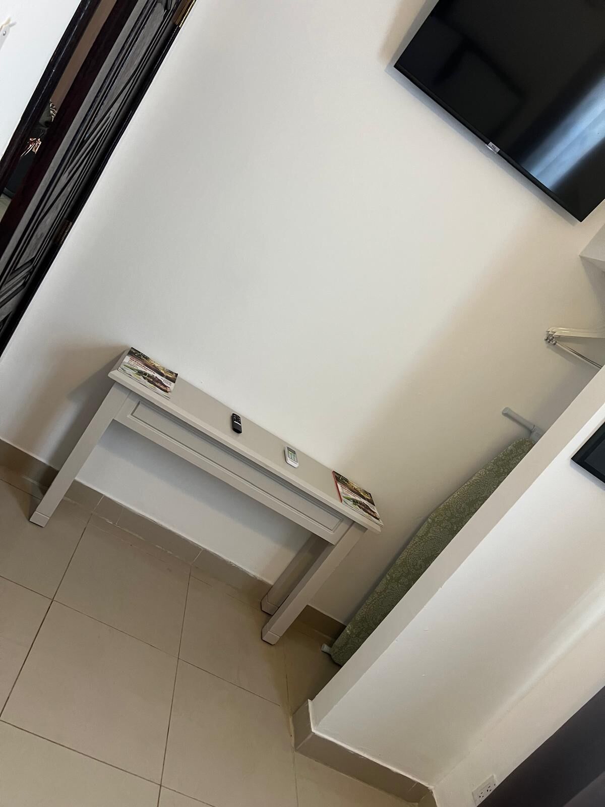 Un rincón de una habitación con un banco blanco, un estante pequeño con un control remoto y un televisor fijado a la pared.