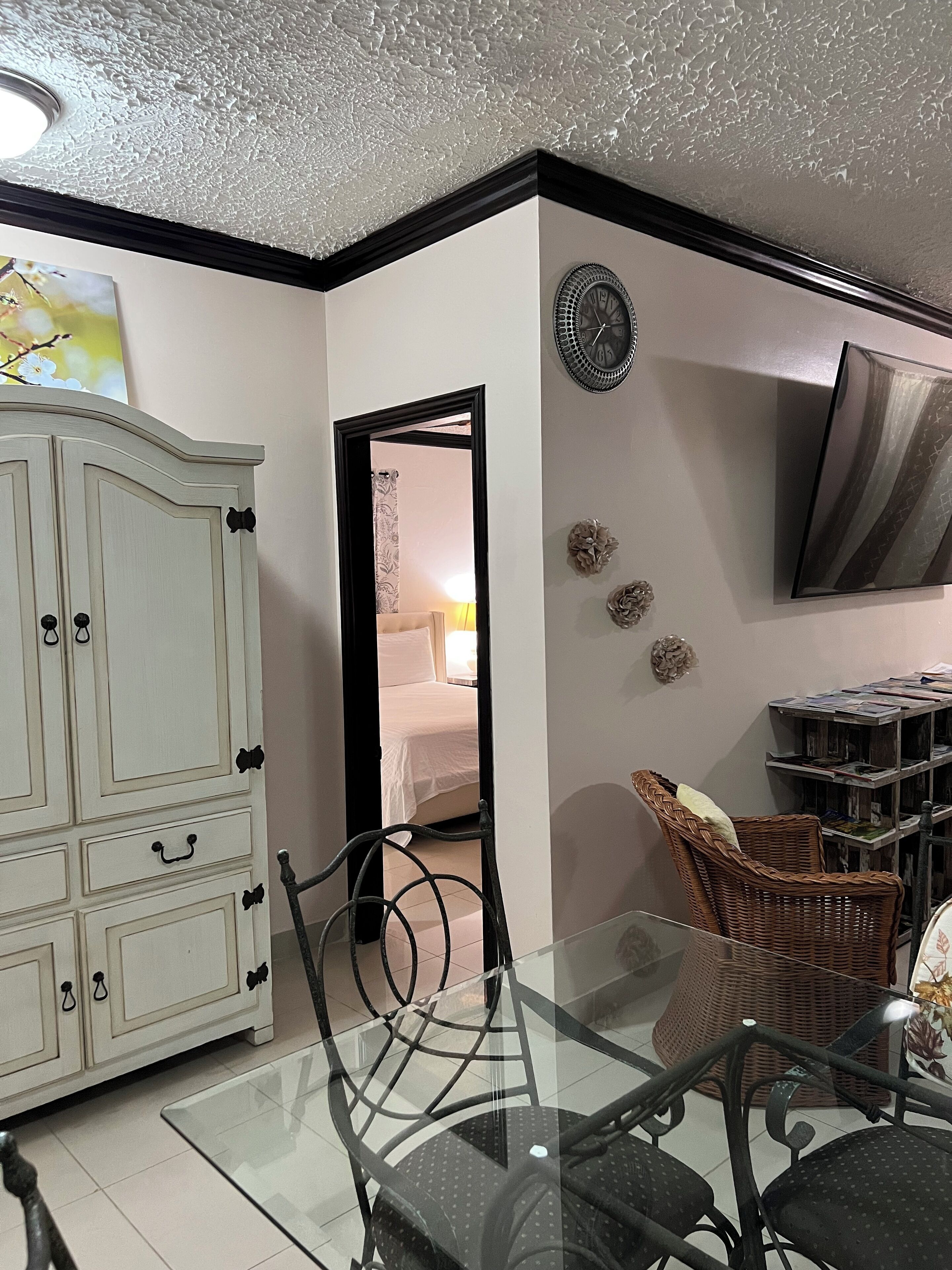 Een kamer met een witte kast, een glazen tafel met stoelen, een rieten stoel, een televisie en een wandklok.