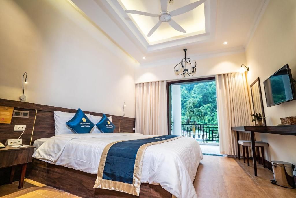 A hotel room with a bed, a desk, a chair, a TV, a ceiling fan, and a view of greenery outside.