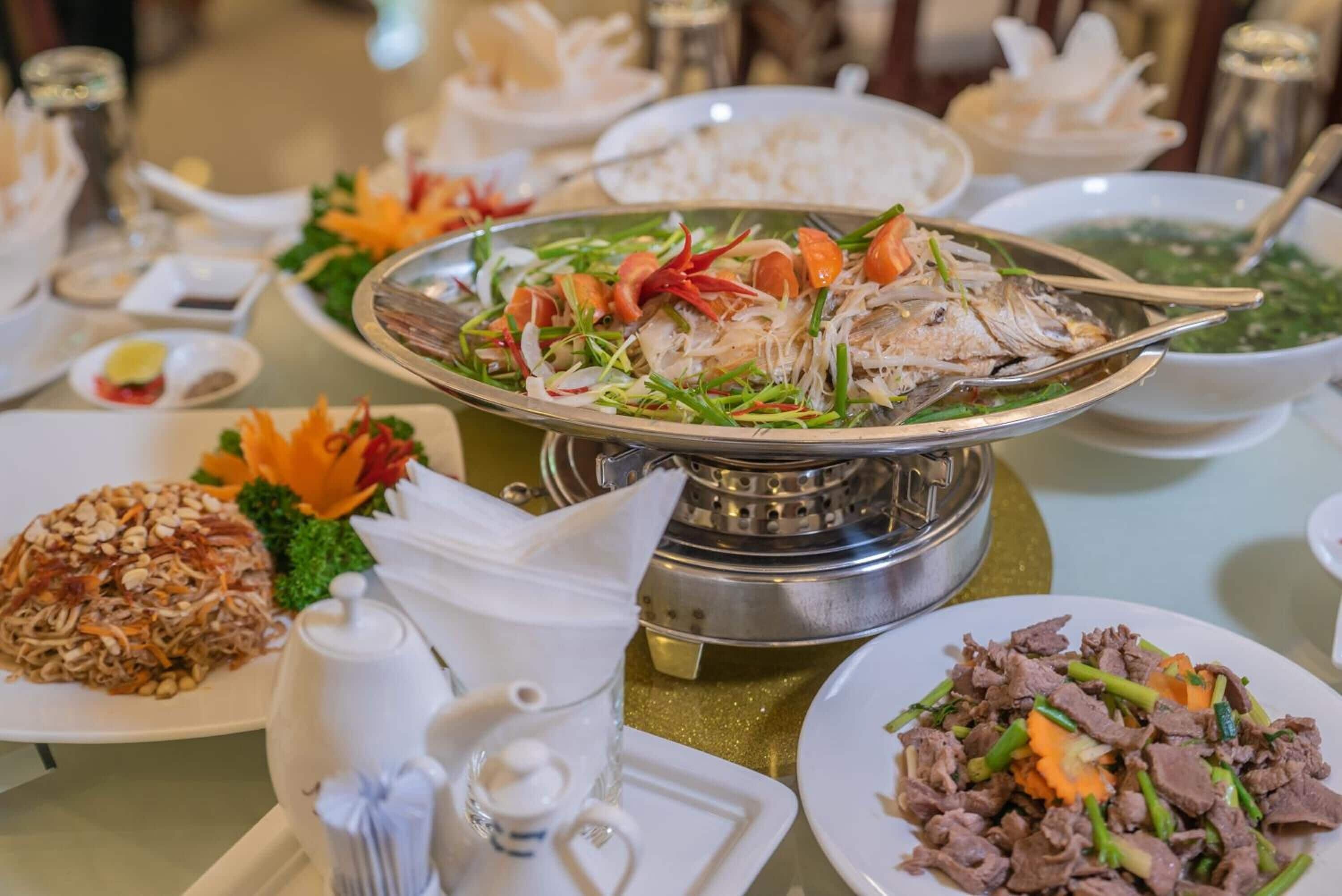 A dining table with various dishes, including a fish dish, a noodle dish, and a beef dish, along with condiments and a teapot.