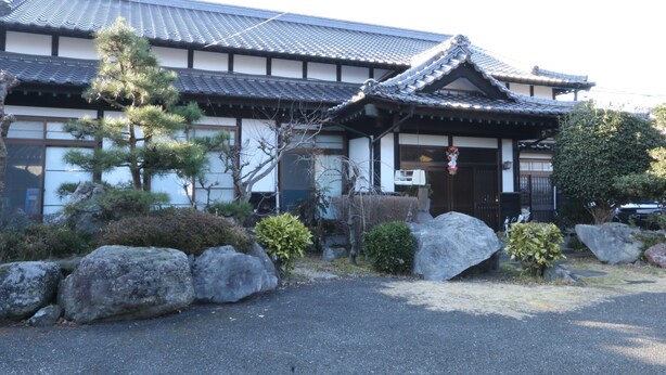 Un edificio japonés tradicional con techo de tejas, rocas grandes y un pino al frente.