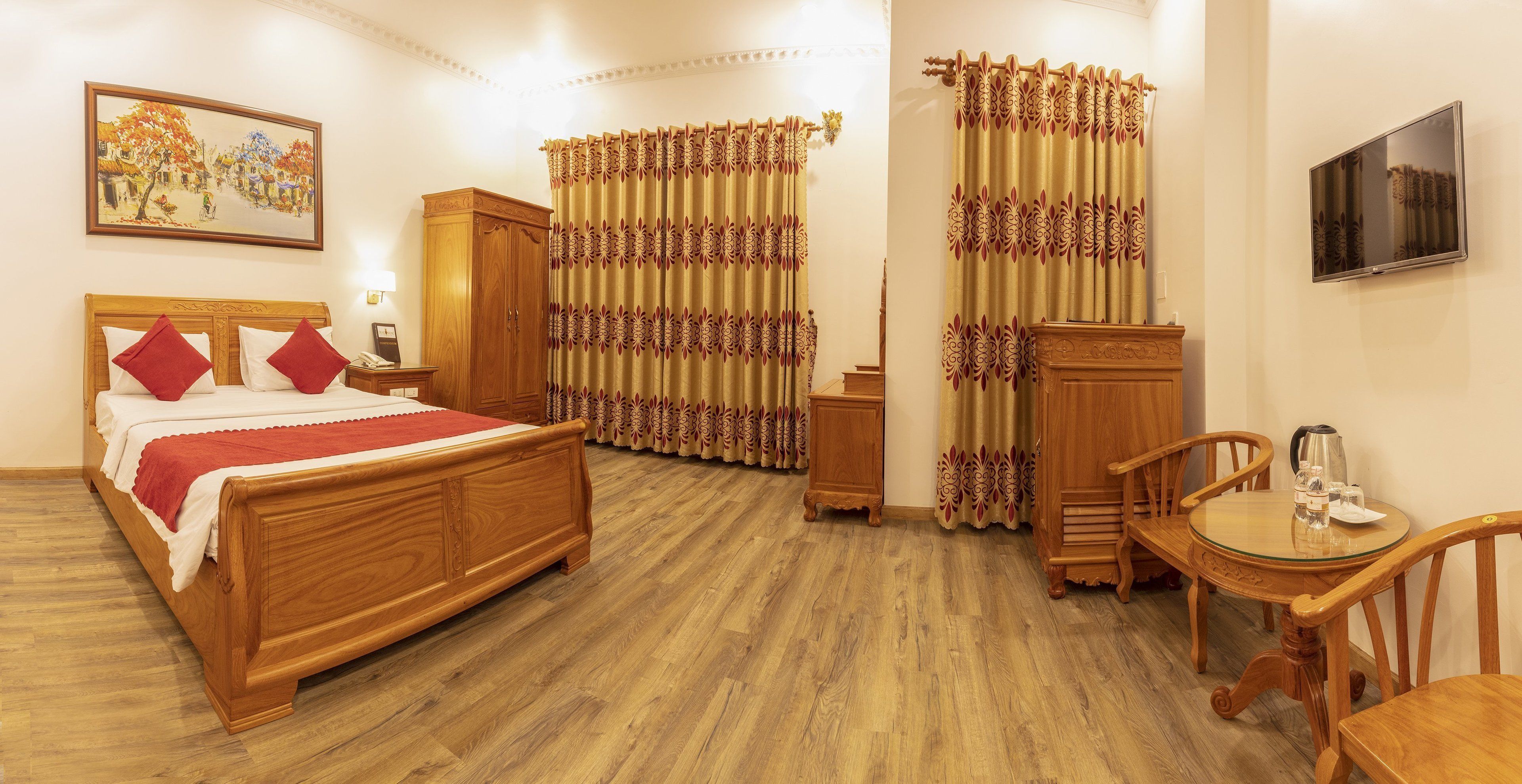 A hotel room with a wooden bed, a small round table with a kettle, a wooden cabinet, and curtains with a floral pattern.