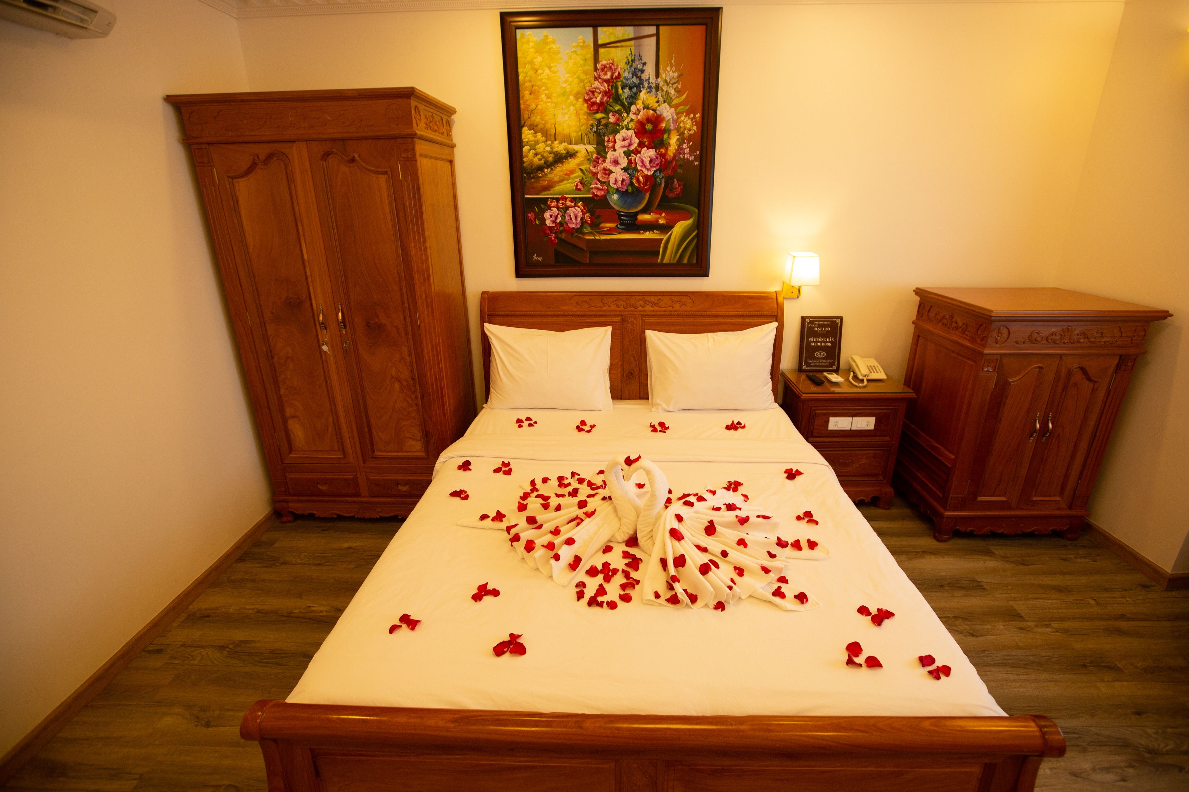 A neatly made bed with a swan and rose petals, wooden furniture, and a painting on the wall.