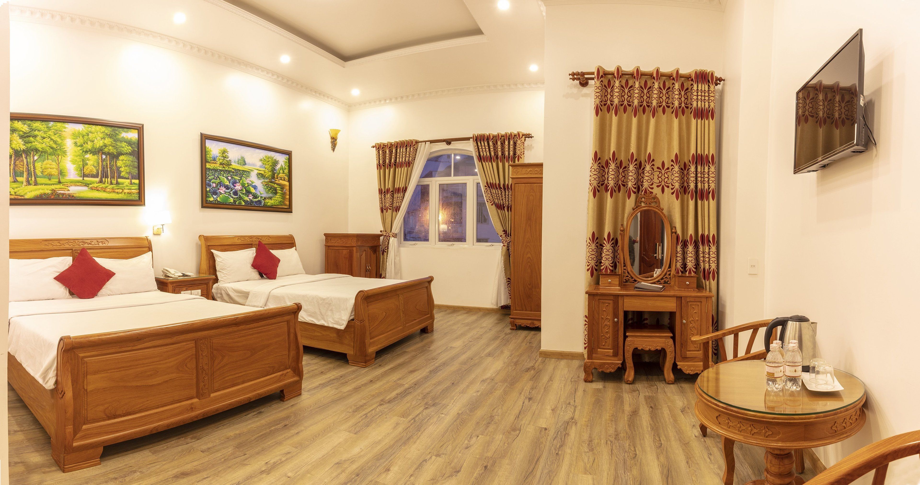 Habitación de hotel con dos camas de madera, un aparador de madera, una mesa redonda con una silla y un televisor montado en la pared.