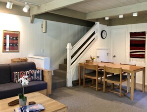 Una sala de estar acogedora con sofá, mesa de centro y una mesa de comedor con sillas. Una escalera conduce a otro nivel.