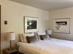 Una habitación de hotel con una cama, dos cuadros en la pared, una mesita de noche con una lámpara y otra mesita junto a la cama.