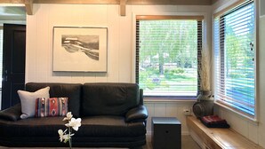 Uma sala de estar com um sofá de couro preto, um quadro na parede, uma janela com persianas e uma mesa pequena com revistas e um vaso de flores.