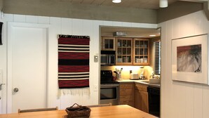 Uma área de refeições com mesa e cadeiras de madeira, uma cozinha com armários e eletrodomésticos, e um quadro fixado na parede.