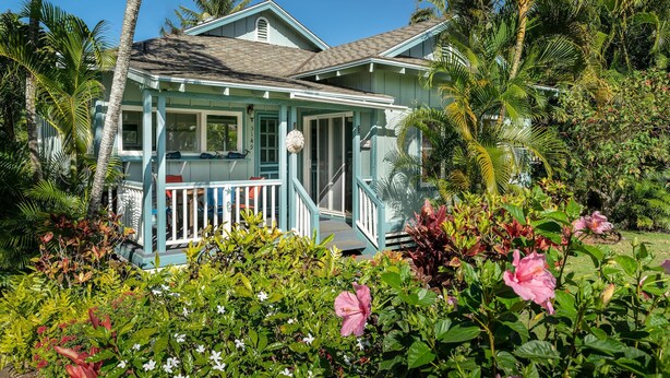 A light blue house with a porch, surrounded by lush tropical plants and flowers.