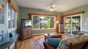 A spacious living room with a television, wooden furniture, a large window with a view of a house and greenery, and a ceiling fan.
