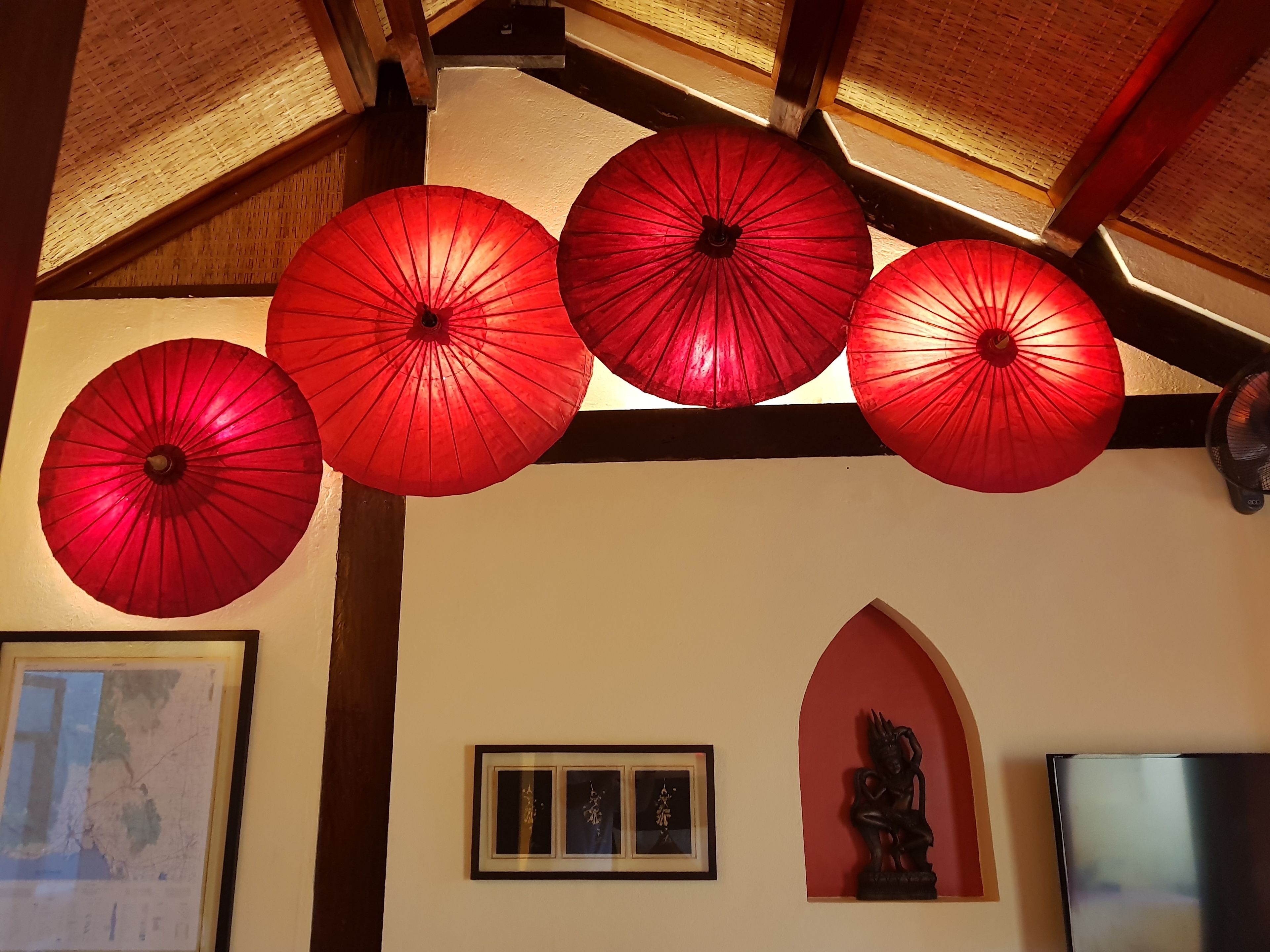 A room with a wooden ceiling, red umbrellas hanging from the ceiling, a framed map, and a statue.
