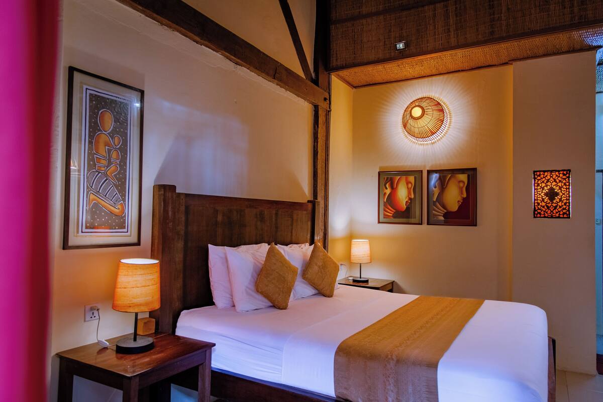 A hotel room with a wooden headboard, a large bed with white linens, two bedside tables with lamps, and artwork on the walls.