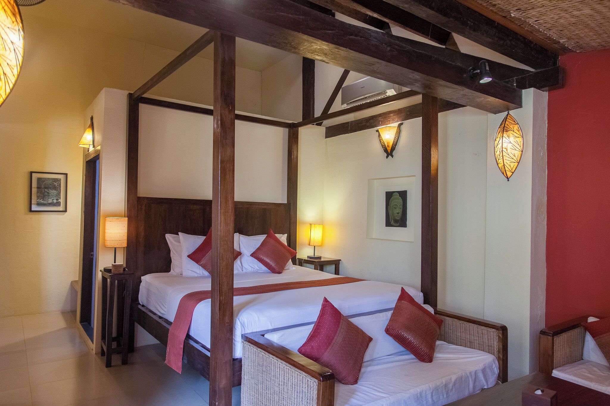 A four-poster bed with white linens and red pillows, wooden headboard, and a nightstand with a lamp.