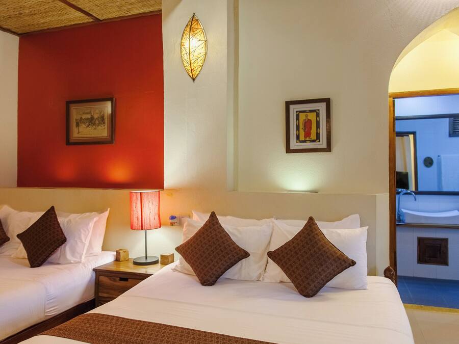 Habitación de hotel con dos camas, una pared de acento roja, un cuadro enmarcado, una lámpara de pared e vista al baño con lavabo y espejo.
