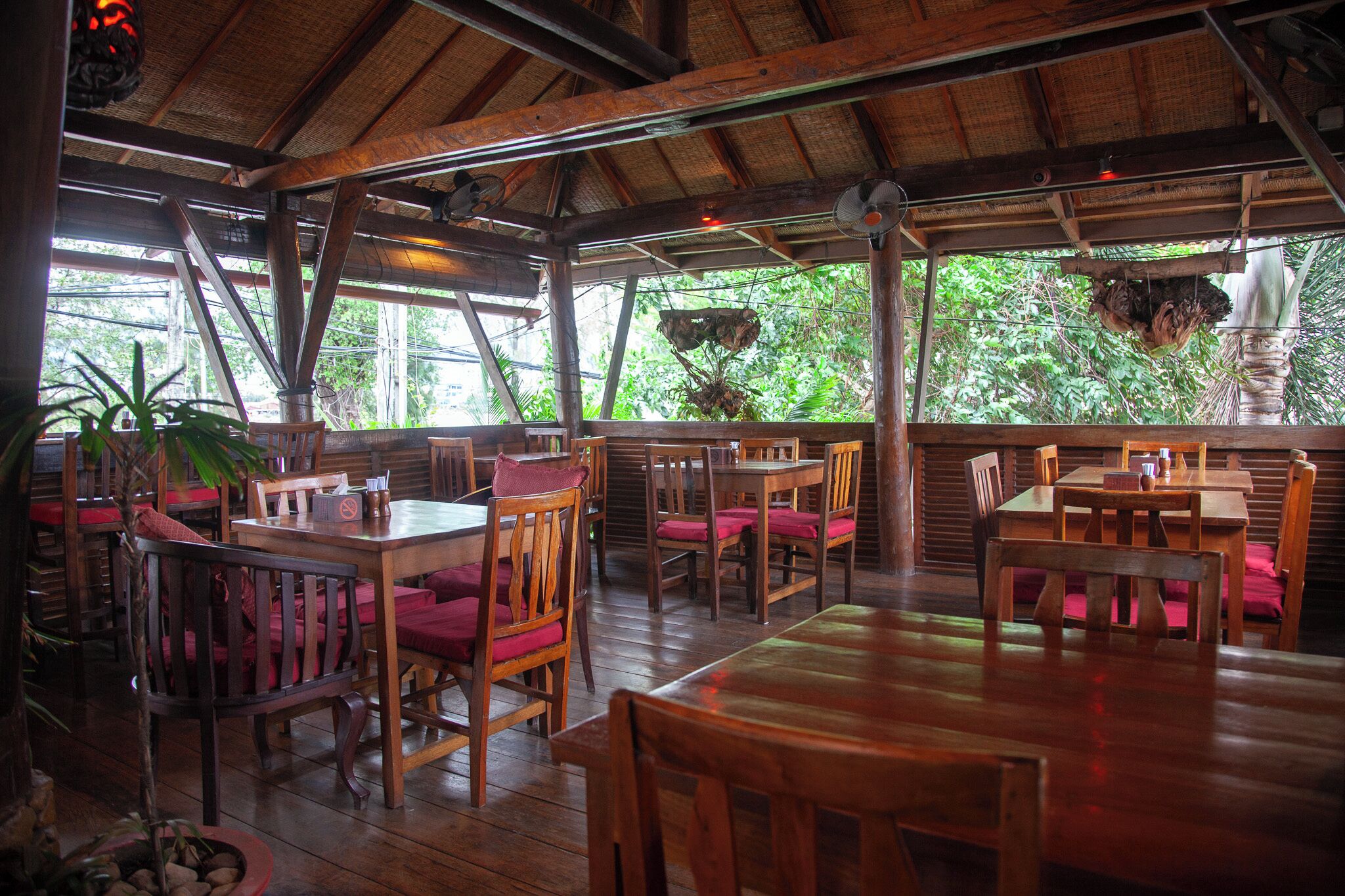 Restaurante com mesas e cadeiras de madeira, telhado de palha e plantas em vasos.
