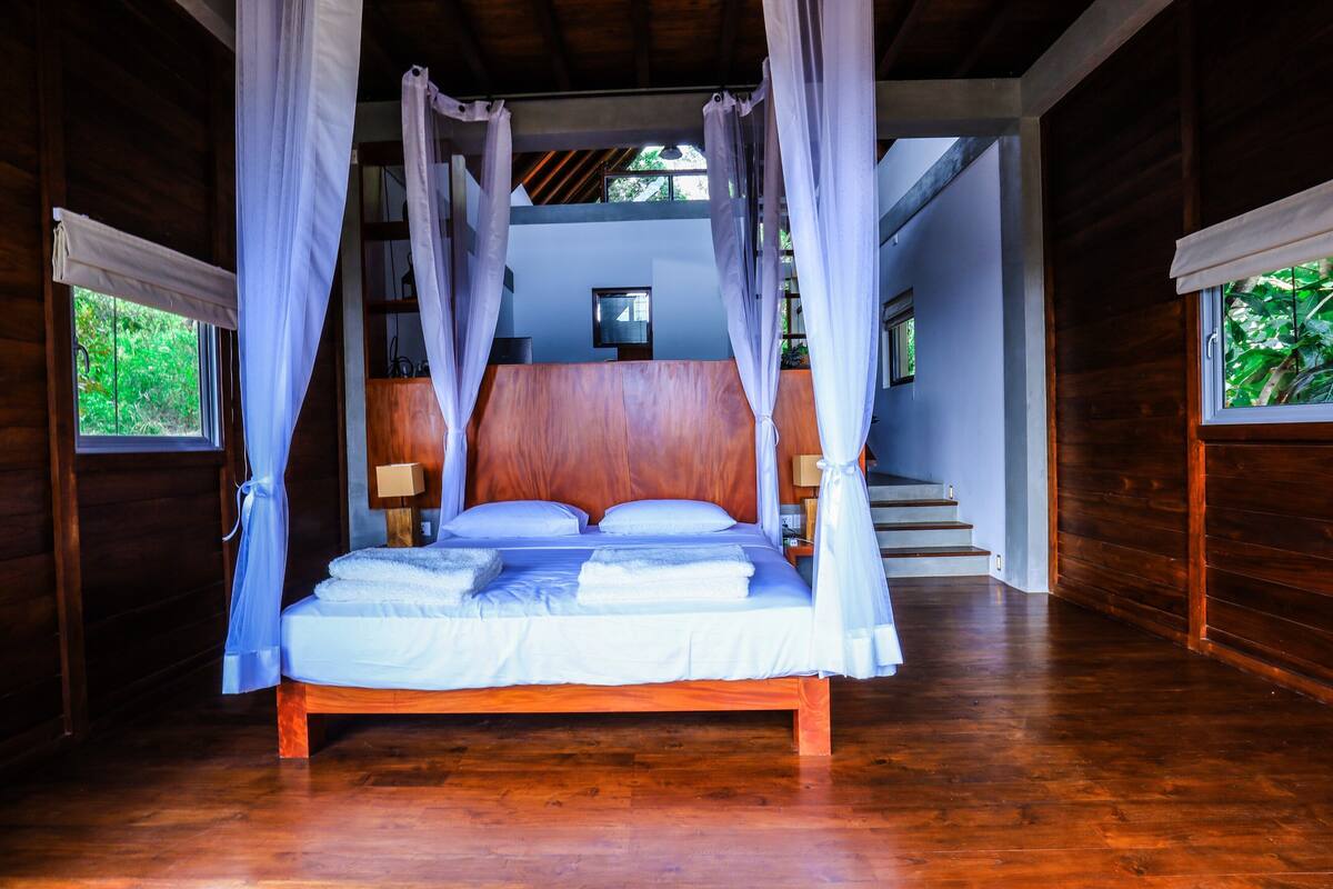 A wooden-framed bed with a canopy, a nightstand with a lamp, and a staircase leading to an upper level.