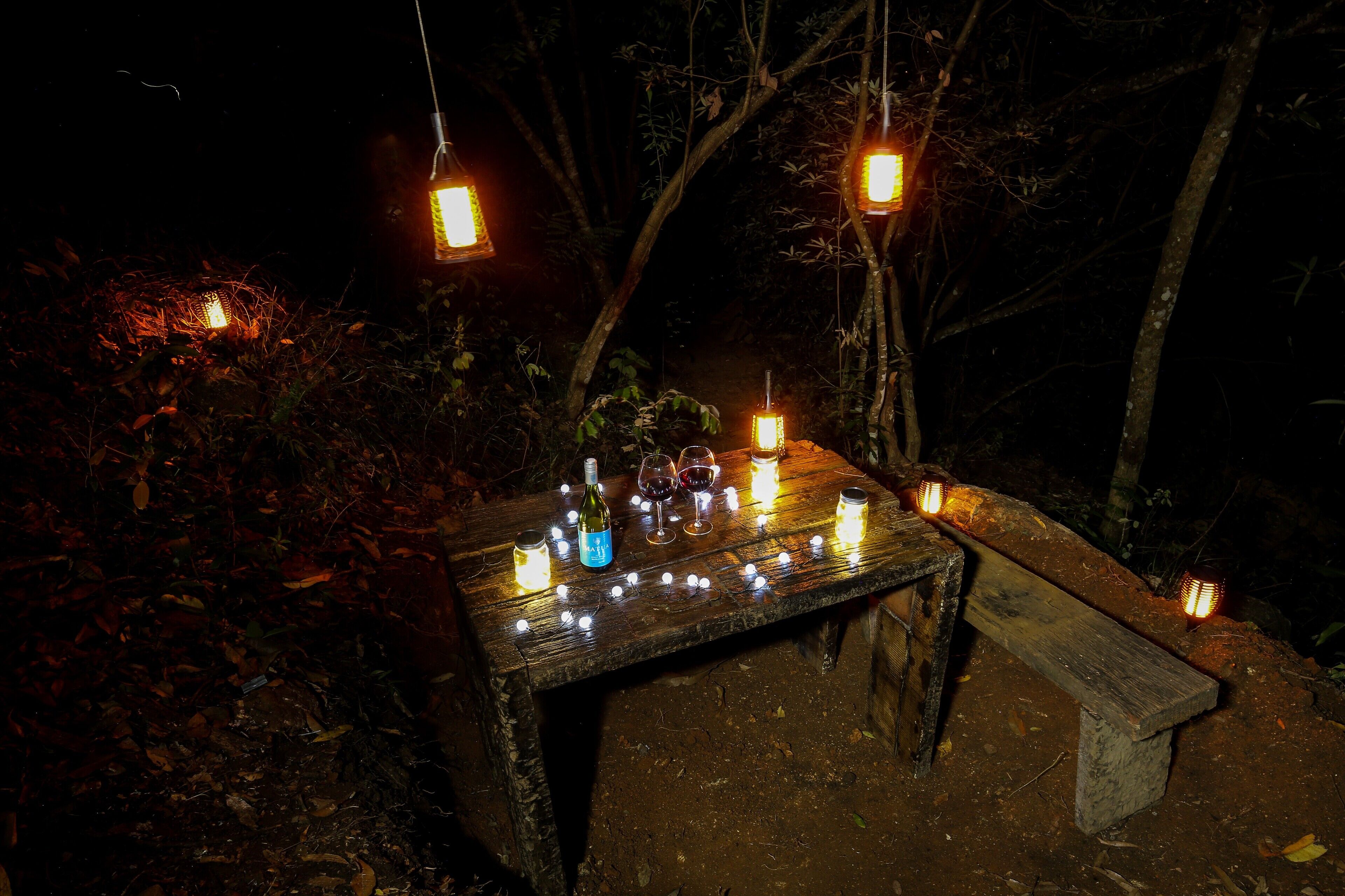 A rustic wooden table with candles and bottles, surrounded by hanging lanterns in a forest setting.