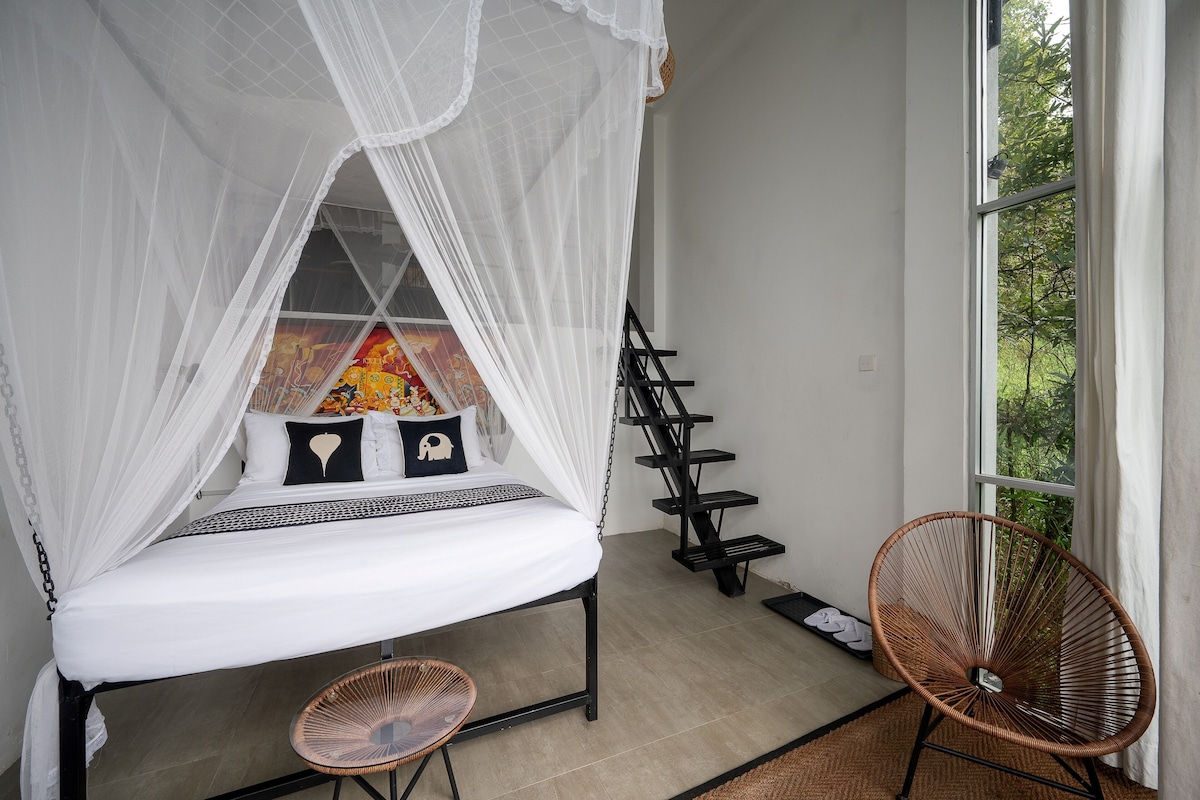 A room with a bed under a white canopy, a chair, a small table, and a window with a view of greenery.