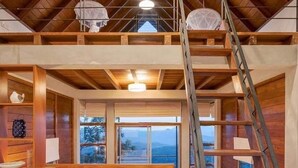 Interior de madera con varios niveles, un escritorio central de madera, dos taburetes, una escalera y una ventana con vistas.