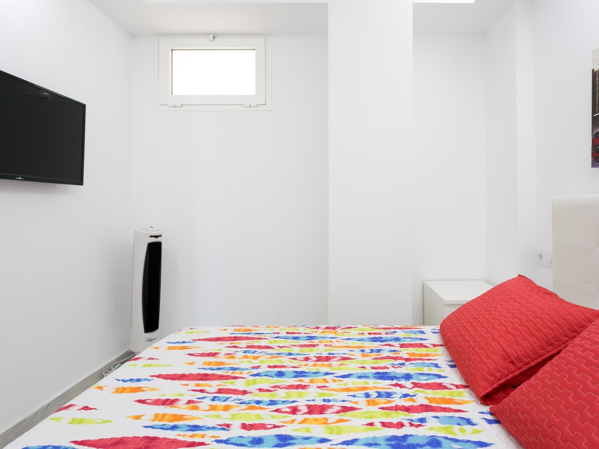 Een kleine, witte kamer met een bed, een raam, een aan de muur bevestigde televisie en een radiator.