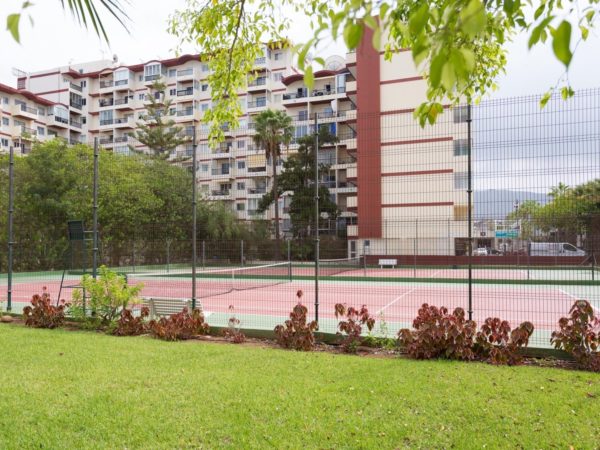 A tennis court surrounded by a fence, with a multi-story residential building in the background.