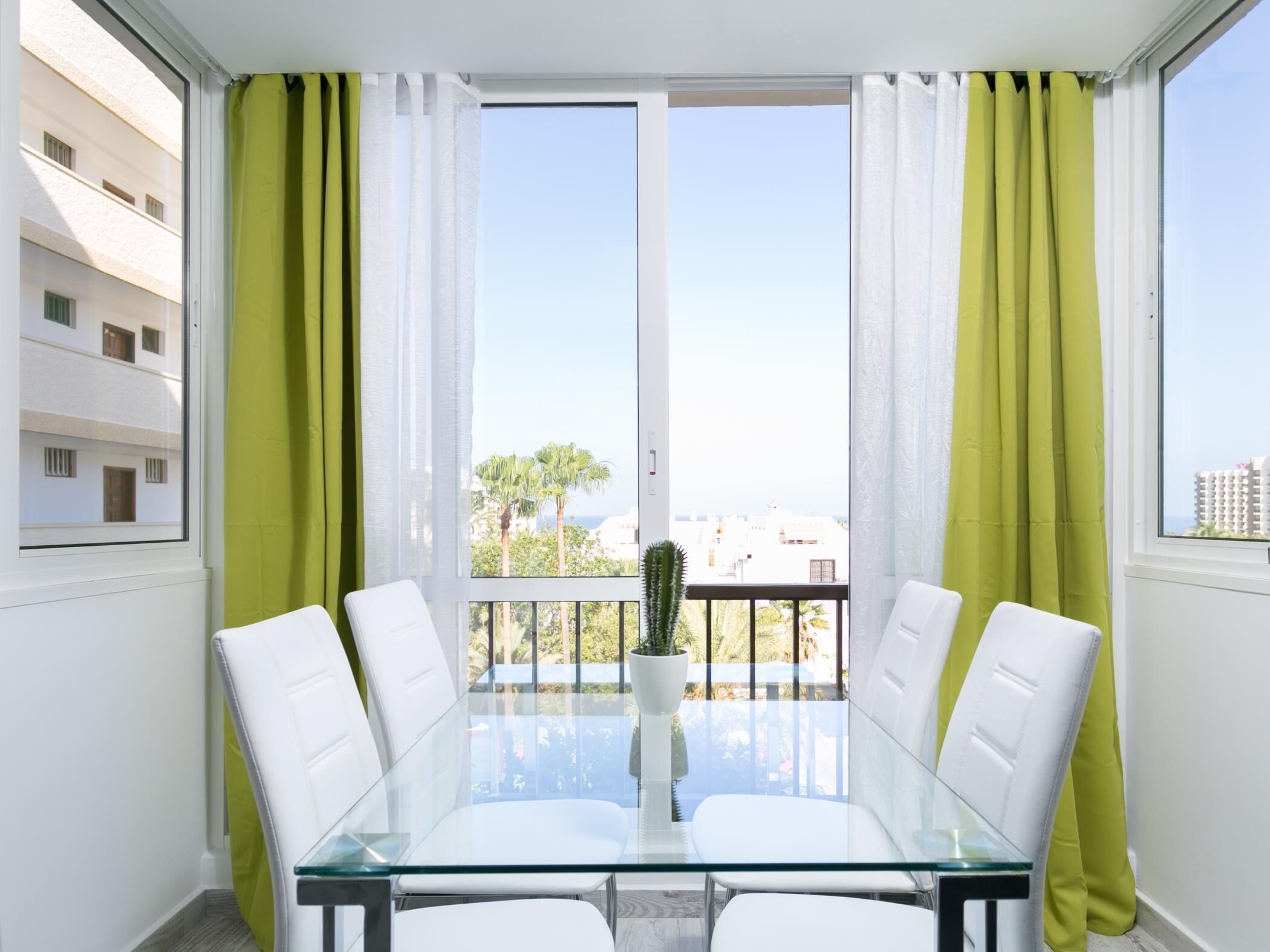 Una habitación moderna con una mesa de vidrio y sillas blancas, ventanas grandes con cortinas verdes y vistas a edificios y palmeras.