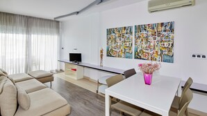A modern living room with a beige sectional sofa, a white dining table with chairs, a flat-screen TV on a stand, and abstract art on the wall. - APARTMENT (5 ADULTS ) POOL VIEW (L'Estartit)