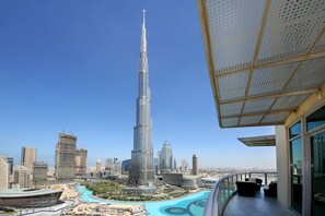 A high-rise building with a distinctive spire, surrounded by other modern skyscrapers, a pool, and a balcony with outdoor furniture.