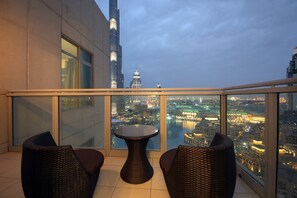 A balcony with wicker furniture and a glass table, overlooking a cityscape with illuminated buildings and a river.