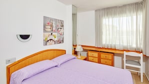 Une chambre à coucher avec un lit, un bureau et une chaise. On y trouve une lampe fixée au mur, un miroir et un cadre contenant diverses photos ou objets.