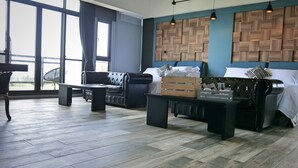 Una habitación de hotel moderna con un ventanal grande, un sofá de cuero negro, una mesa de centro de madera y una cama con cabecera azul.