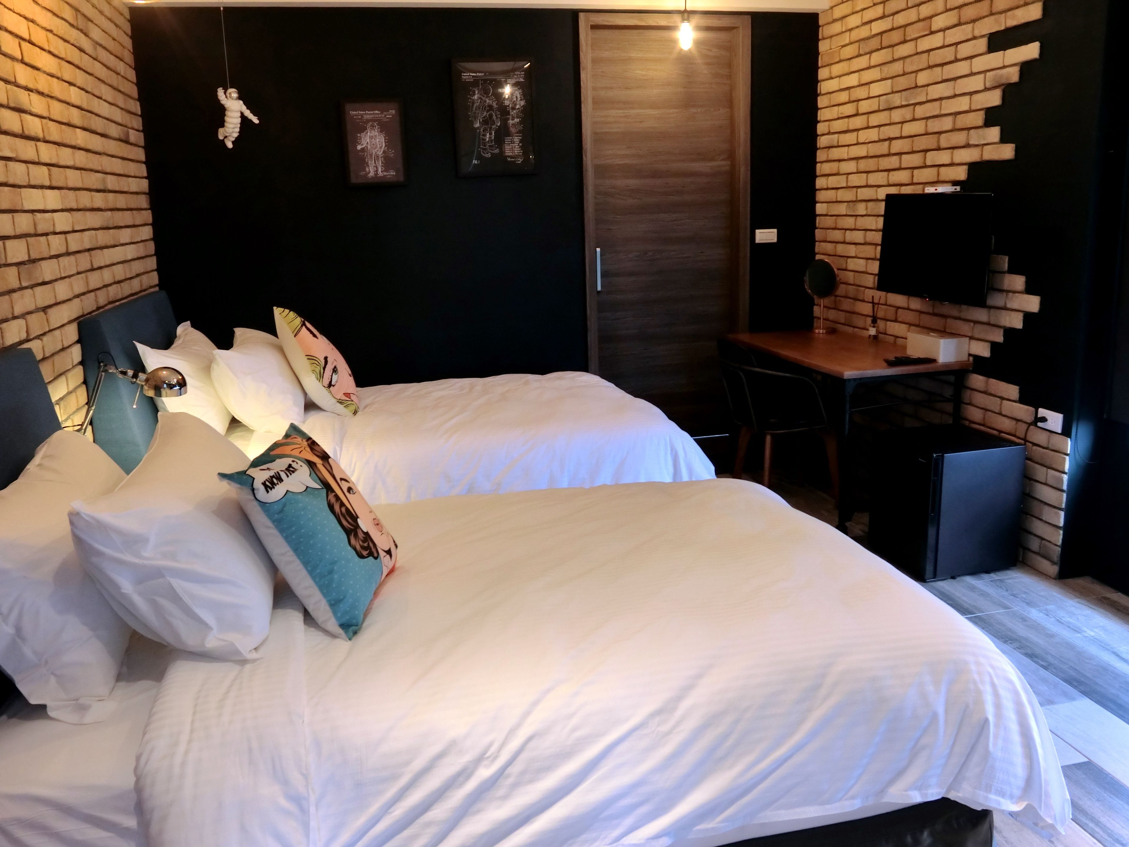 A hotel room with two beds, a desk, and a brick wall.