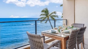 A balcony with a view of the ocean, a table set for dining, wicker chairs, and a palm tree in the background. - Tranquil OceanFront 2BD AC Pool, Amazing Views - Mahana 519 (Lahaina)