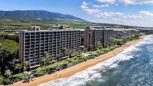 A hotel with multiple floors, a beachside location, and a view of mountains in the background. - Tranquil OceanFront 2BD AC Pool, Amazing Views - Mahana 519 (Lahaina)